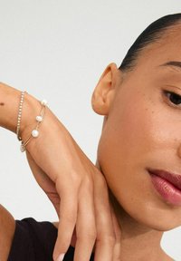 Pulsera de oro con perlas y pedrería, con un delicado diseño de cadena. El tono de piel suave y el fondo neutro realzan la joyería.