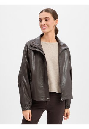 Femme portant une veste en cuir marron foncé sur un pull beige et un pantalon foncé, souriante et regardant sur le côté sur un fond uni.