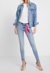 Chaqueta de mezclilla azul claro sobre una blusa blanca, combinada con jeans skinny azul claro desgastados. Bufanda rosa anudada en la cintura, tacones color nude.