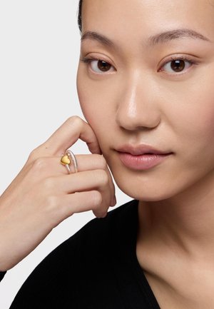 Close-up of a person with clear skin wearing a silver ring with a small gold heart, resting their hand against their face, wearing a black top.