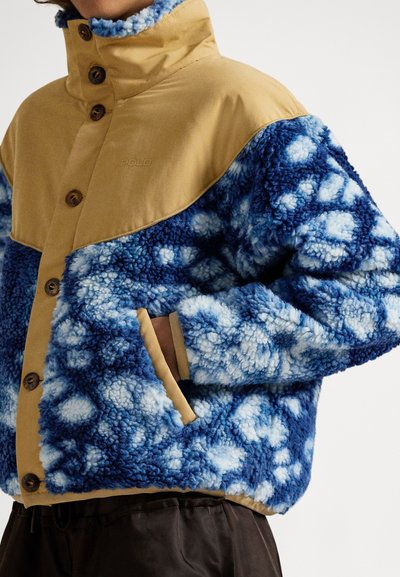 Ζακέτα από φλις με ταμπά επάνω μέρος, μπλε υφή στο κάτω μισό, κουμπιά στο μπροστινό μέρος και δύο πλαϊνές τσέπες. Το σχέδιο περιλαμβάνει ανοιχτό μπλε στίγματα.