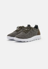 Sneakers sportive con una tomaia in tessuto grigio texturizzato, dettagli in verde oliva e suola in gomma bianca. Presentano lacci tondi e un'etichetta per il sollevamento.