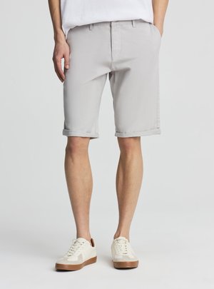 Uomo che indossa pantaloncini grigio chiaro con orlo risvoltato e tasche laterali e scarpe da ginnastica bianche con suole marroni, in piedi davanti a uno sfondo semplice.