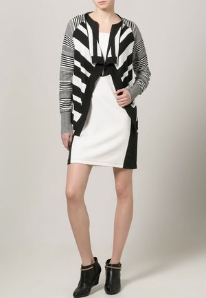 Czarny i biały kardigan w paski noszony na białej sukience, z kontrastowymi czarnymi akcentami. Botki zdobione złotym łańcuszkiem. Gładka tekstura.