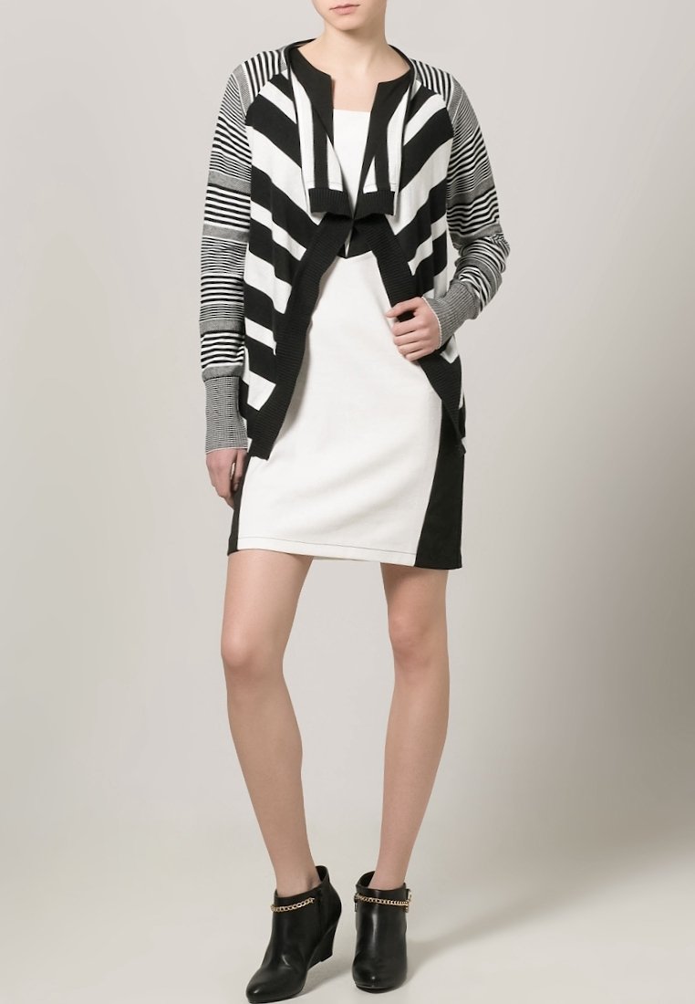 Cárdigan de rayas en blanco y negro sobre un vestido blanco, con acentos negros en contraste. Botines con detalles de cadena dorada. Textura suave.