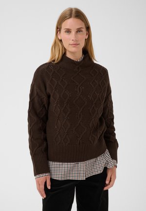 Femme portant un pull en maille torsadée marron par-dessus une chemise à carreaux, debout devant un fond clair uni.