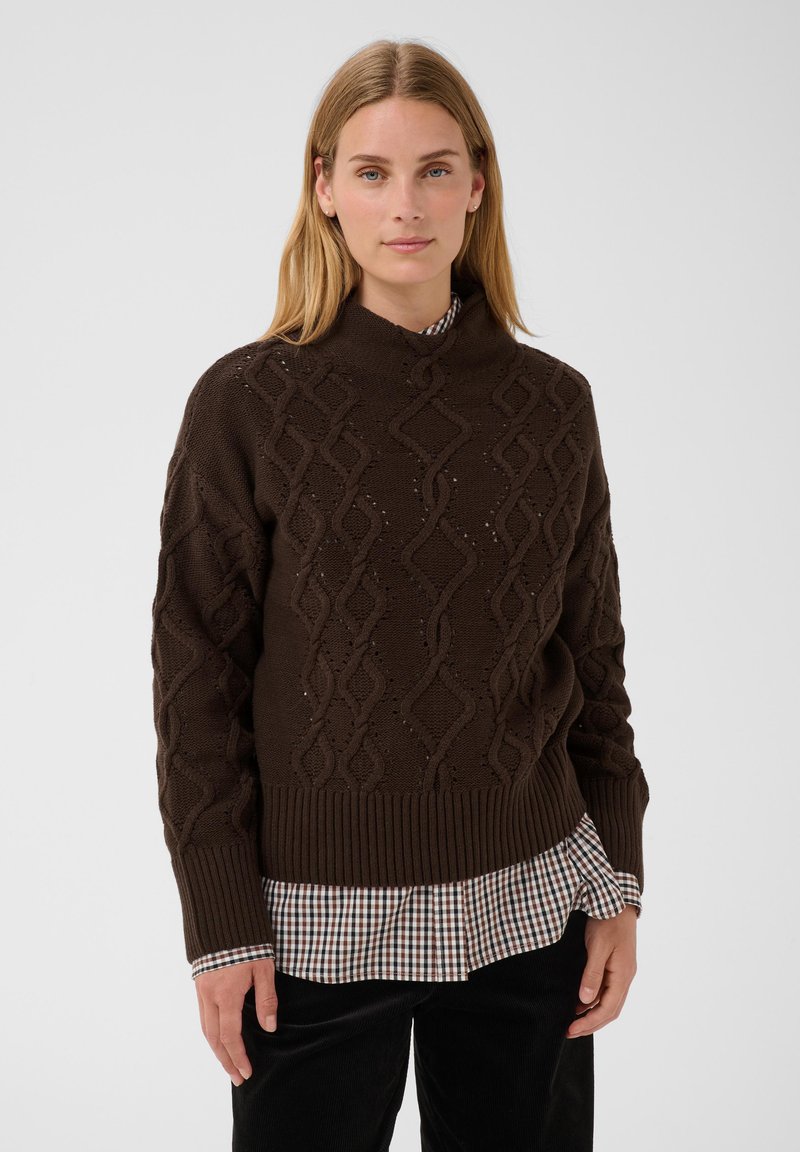 Femme portant un pull en maille torsadée marron par-dessus une chemise à carreaux, debout devant un fond clair uni.