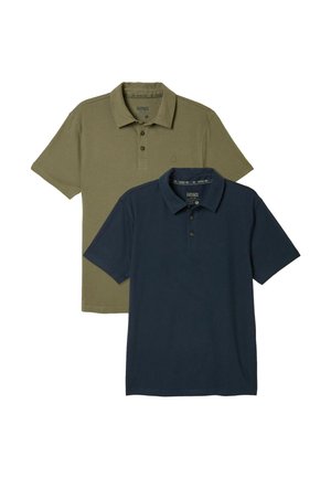 Man met baard in olijfgroene polo met korte mouwen en lichtblauwe spijkerbroek, staand tegen een effen witte achtergrond.