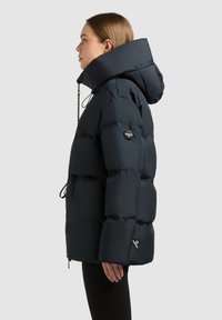 Navy blue puffer jacket with a high collar, large hood, and adjustable drawstrings. Features a logo patch and side zip pockets.