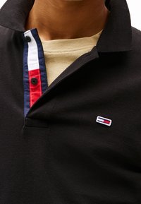 Svart polo skjorta med texturerad tyg, med en flerfärgad krage i rött, vitt och blått och en liten logotyp på bröstet.