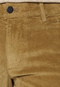 Corduroybroek in een warme tan kleur, met verticale ribbels, een zwarte knoop en klassieke stikdetails langs de naden.