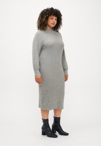 Vestido de malha em cinza claro com textura canelada, decote redondo e mangas longas. Combinado com botas de tornozelo pretas com salto quadrado.