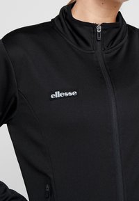 Schwarze Zip-Jacke aus glattem Stoff, mit Stehkragen, seitlichen Taschen und einem gestickten "ellesse"-Logo auf der Brust.