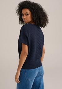 Tumsā zila adīta džemperis ar īsām piedurknēm, ar zīmolposma rakstu un ribotu apdari. Apvienots ar gaiši ziliem džinsiem.