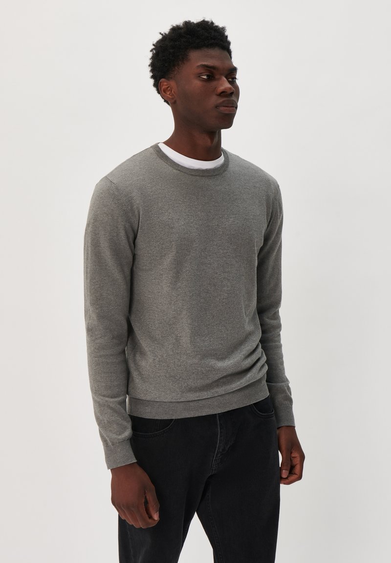 Suéter gris de punto con cuello redondo y mangas largas, que presenta un cuello y puños acanalados, combinado con pantalones oscuros. Diseño minimalista.