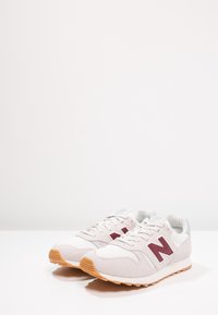 Baskets blanches avec un logo "N" gris clair et bordeaux, en suède et mesh, semelle extérieure en caoutchouc, et design à lacets.