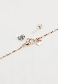 Bracelet en chaîne en or rose avec un fermoir à ressort, comportant un charm en forme de logo circulaire et deux clous d'oreilles assortis dans diverses teintes métalliques.
