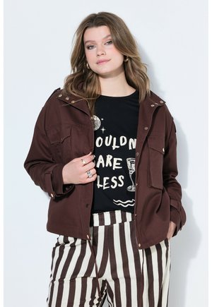 Junge Frau trägt eine braune Jacke, ein schwarzes T-Shirt mit Grafik und weiße und braun gestreifte Hosen mit vertikalen Streifen, posiert vor einem weißen Hintergrund.