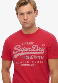 Rood katoenen T-shirt met een grote grijze grafische print met "SuperDry" en aanvullende tekst in meerdere talen. Klassiek crew neck ontwerp.