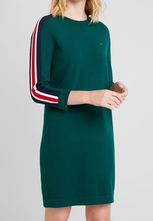 Femme portant une robe verte à manches longues avec des rayures rouges, blanches et bleu marine le long des manches, debout devant un fond gris uni.