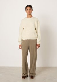 Kremas, purus megztinis su rilės tekstūra, derinamas su laisvomis alyvuogių žaliomis kelnėmis; modelis stovi priešais paprastą foną.