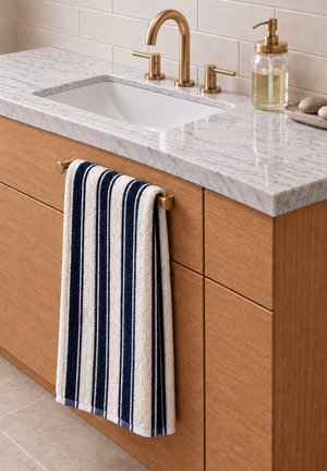 Lavabo de salle de bain avec robinet en laiton, plan de travail en marbre blanc, serviette rayée bleu marine et blanche sur un meuble en bois, et distributeur de savon transparent avec savon liquide.