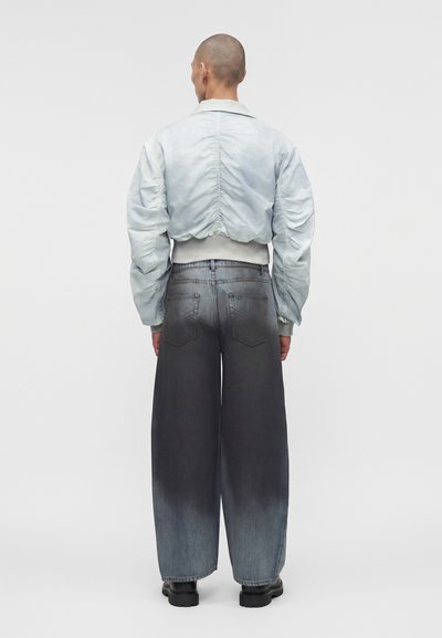 Άτομο με ξυρισμένο κεφάλι που φοράει ένα ανοιχτό μπλε μπουφάν bomber με πιέτες και φαρδιά τζιν με σκούρες αποχρώσεις, στέκεται αντιμετωπίζοντας ένα απλό ανοιχτό γκρι φόντο.