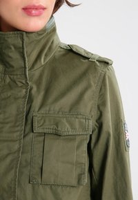 Kurtka wojskowa w oliwkowej zieleni z wysokim kołnierzem, dwoma przednimi kieszeniami, pagonami na ramionach oraz naszywką na rękawie.
