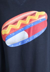 Camiseta azul marino con un gráfico de un perrito caliente con mostaza amarilla, acentos rojos y azules, y el texto "TOMMY JEANS" en blanco.