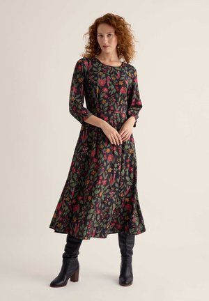 Femme aux cheveux roux bouclés portant une robe noire fleurie à manches trois-quarts et des bottines noires à talons, debout sur un fond uni.