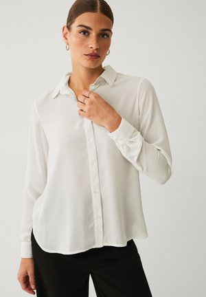 Vrouw met naar achteren gebonden haar, zilveren oorringen, een doorschijnende witte blouse met knopen en een zwarte broek, haar blouse bij de kraag aanrakend.