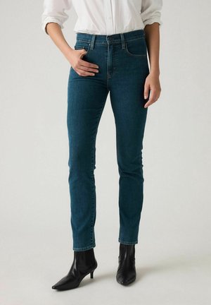 Personne portant un jean skinny bleu foncé, une chemise blanche boutonnée et des bottines noires à bout pointu, debout devant un fond uni.