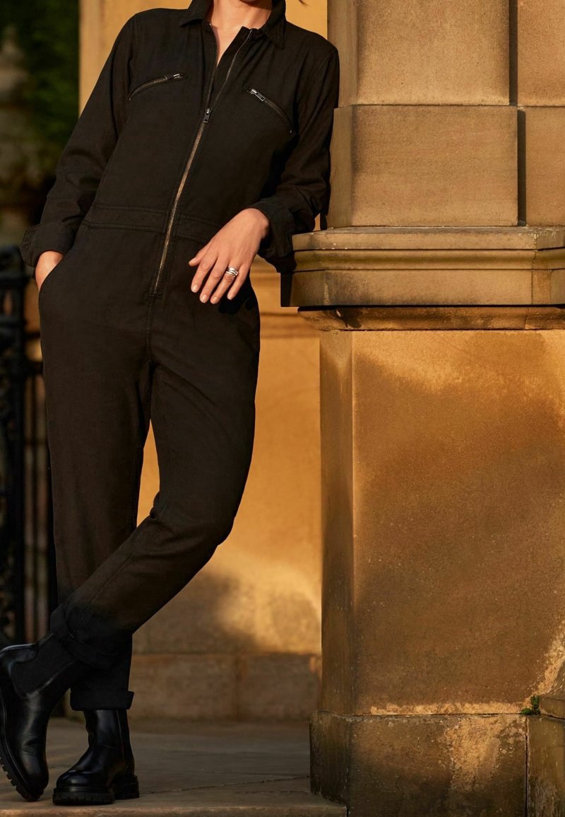 Zwarte jumpsuit met een ritssluiting aan de voorkant, lange mouwen en twee borstzakken; gecombineerd met zwarte laarzen, tegen een stenen muur.