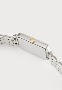Montre en métal argenté avec un boîtier rectangulaire, couronne dorée et bracelet en acier inoxydable présentant des maillons entrelacés. Fini lisse et poli.
