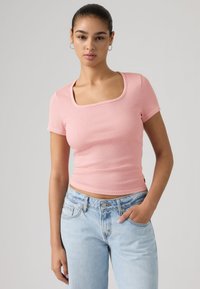 Νεαρή γυναίκα με σκούρα μαλλιά, φορώντας ροζ κοντομάνικο πλεκτό τοπ και ανοιχτόχρωμο μπλε τζιν, στάζει με το ένα χέρι στην τσέπη, μπροστά σε ουδέτερο φόντο.