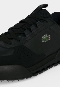 Baskets en cuir noir avec lacets noirs, détails perforés et petit logo crocodile vert sur le côté.