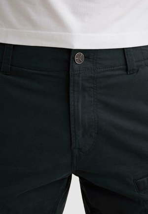 Detailný záber na tmavozelené nohavice s kovovým gombíkom a zipsom na prednom diely, nosené s bielou košeľou zastrčenou dovnútra.