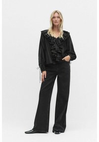 Svart volangblus med långa ärmar och dragsko-detaljer, matchad med högmidjade vida svarta jeans med en subtil textur.