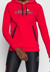 Sweat à capuche rouge avec cordons noirs et logo "ellesse", porté avec des leggings de sport noirs ornés de textes blancs et orange.