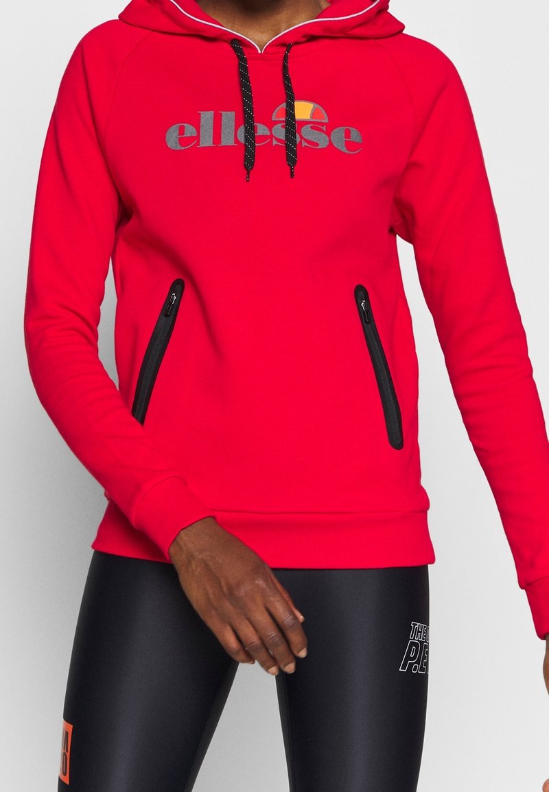 Sweat à capuche rouge avec cordons noirs et logo "ellesse", porté avec des leggings de sport noirs ornés de textes blancs et orange.
