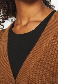 Brun stickad kofta med ribbat mönster, lagd över en svart, åtsittande topp. Har en bred krage och subtila sömnadsdetaljer.