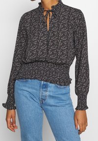 Blusa nera con stampa floreale, vita smock, polsini arricciati e colletto a fiocco. Abbinata a jeans in denim blu chiaro.