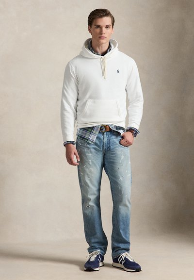 Hvit hettegenser med frontlomme, kombinert med lyseblå slitte jeans. Rutete skjorte synlig ved midjen, komplettert med blå joggesko.