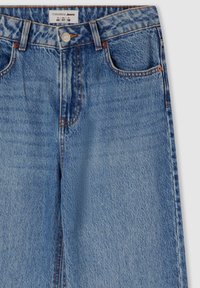 Jeans in denim blu chiaro con vita alta, cinque tasche e rivetti in rame. Presentano una texture sbiadita e una chiusura con bottone sul davanti.