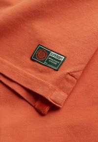 Pomarańczowa, teksturowana tkanina z metką Superdry, z zielonymi i czerwonymi akcentami, naszywką oraz małą pętelką po boku.