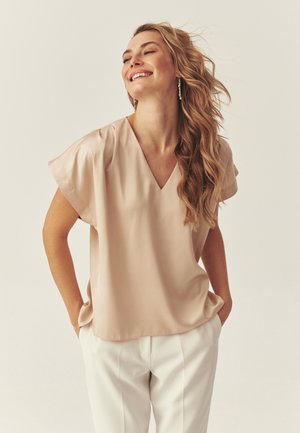 Uśmiechnięta kobieta z długimi falującymi włosami, ubrana w beżową bluzkę z dekoltem w serek i białe spodnie, ręce w kieszeniach, na tle jednolitego jasnego tła.