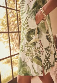 Main d'une femme avec un bracelet chaîne en or dans la poche d'une robe crème à imprimé feuilles tropicales vertes, près d'une fenêtre avec lumière naturelle.