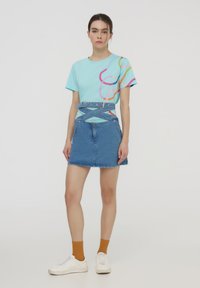 T-shirt azzurro chiaro con motivi astratti multicolore, abbinato a una gonna di jeans con una cintura e una tasca frontale. Sneakers bianche e calzini arancioni.