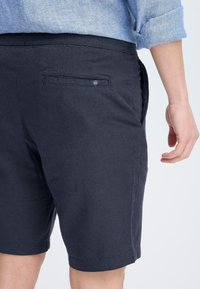 Marineblå shorts laget av lett materiale, med en baklomme med klaff og kontrasterende søm. Sømmer med hemmet kanter, minimal tekstur.