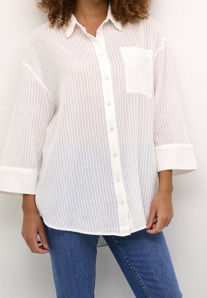 Kvinde iført en halvgennemsigtig hvid skjorte med lodrette striber og blå jeans, stående foran en ensfarvet lys baggrund.